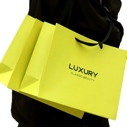 Special Craft Paper Bags feature eye catching fluorescent yellow surface printed with bold black LUXURY CLASSIC BEAUTY lettering. Matching black ribbon handles are smooth and sturdy for comfortable carrying. Thick rigid craft paper keeps the bag upright and holds shape well. The vivid high contrast design delivers strong visual impact and highlights brand style. We support custom bag size base color printed text and brand logo. These paper bags are ideal for fashion stores cosmetic boutiques accessory shops and trendy retail gift packaging.